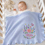 May include: A light blue knitted baby blanket with a ruffled border, featuring an embroidered floral arrangement with a butterfly and the name Mila in pink script. The blanket is draped over a sleeping infant in a white crib with a plush t
