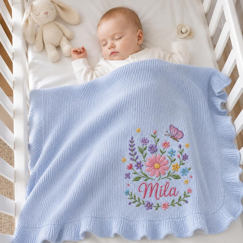 May include: A light blue knitted baby blanket with a ruffled border, featuring an embroidered floral arrangement with a butterfly and the name Mila in pink script. The blanket is draped over a sleeping infant in a white crib with a plush t