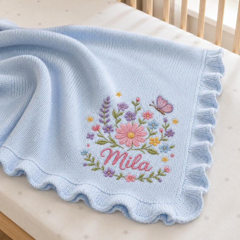 May include: A light blue knitted baby blanket with a ruffled edge. The blanket features an embroidered design of a pink daisy, purple and yellow wildflowers, green leaves, and a small butterfly. The name Mila is embroidered in pink cursive