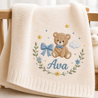 May include: A cream-colored knit baby blanket featuring an embroidered design of a brown teddy bear wearing a blue bow tie. The bear is surrounded by a floral wreath, a blue bow, yellow stars, and a cloud, with the name Ava embroidered in 