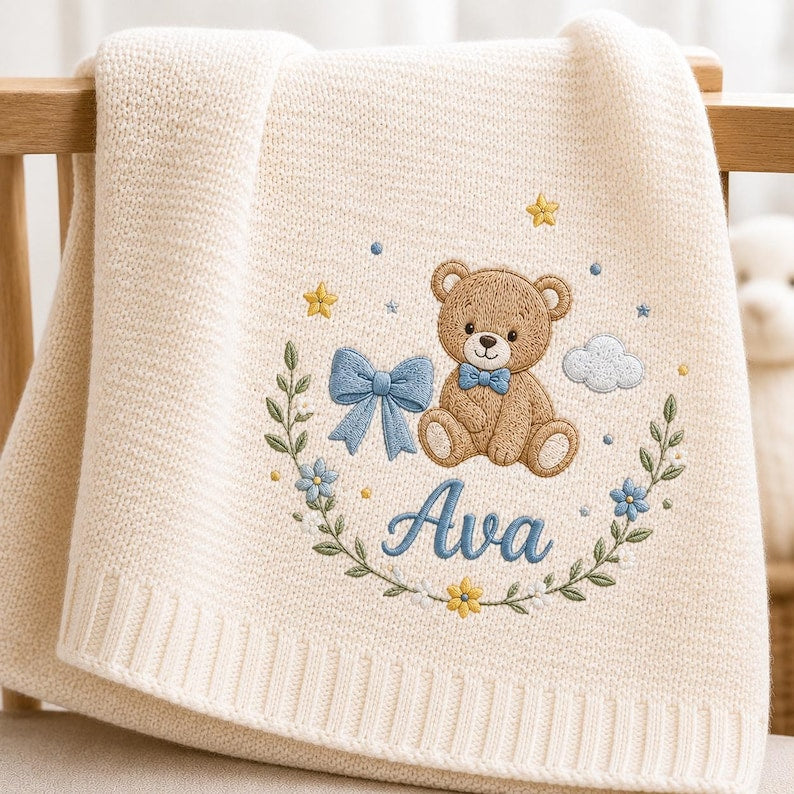 May include: A cream-colored knit baby blanket featuring an embroidered design of a brown teddy bear wearing a blue bow tie. The bear is surrounded by a floral wreath, a blue bow, yellow stars, and a cloud, with the name Ava embroidered in 