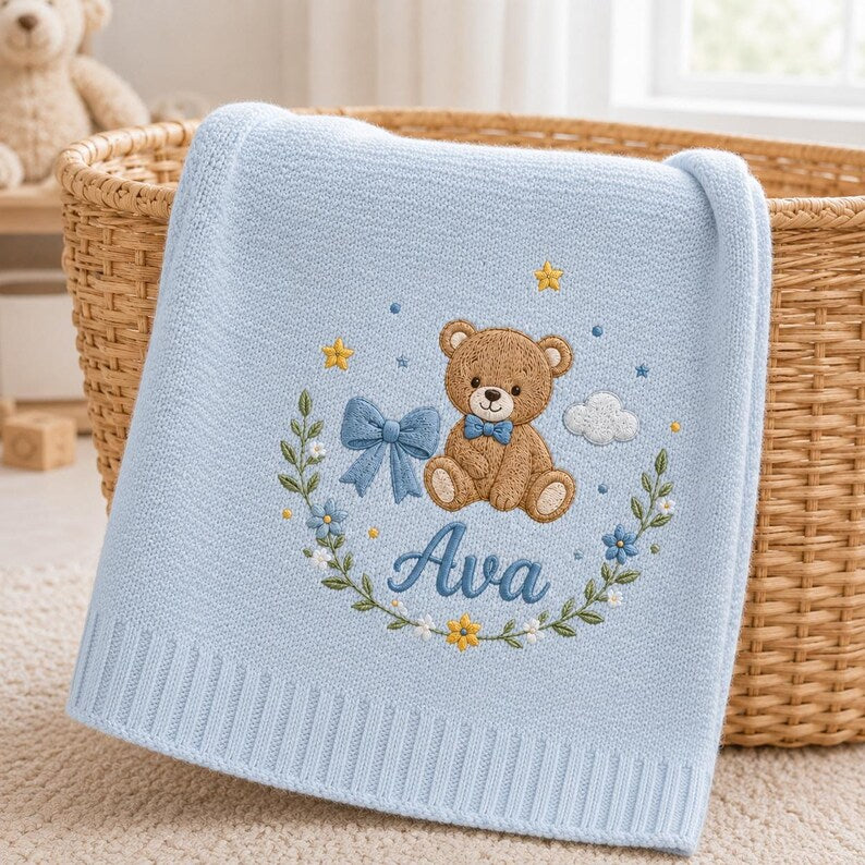 May include: A light blue knitted baby blanket featuring an embroidered brown teddy bear wearing a blue bow tie. The design includes a blue bow, yellow stars, a white cloud, and a floral wreath surrounding the embroidered name Ava in blue s