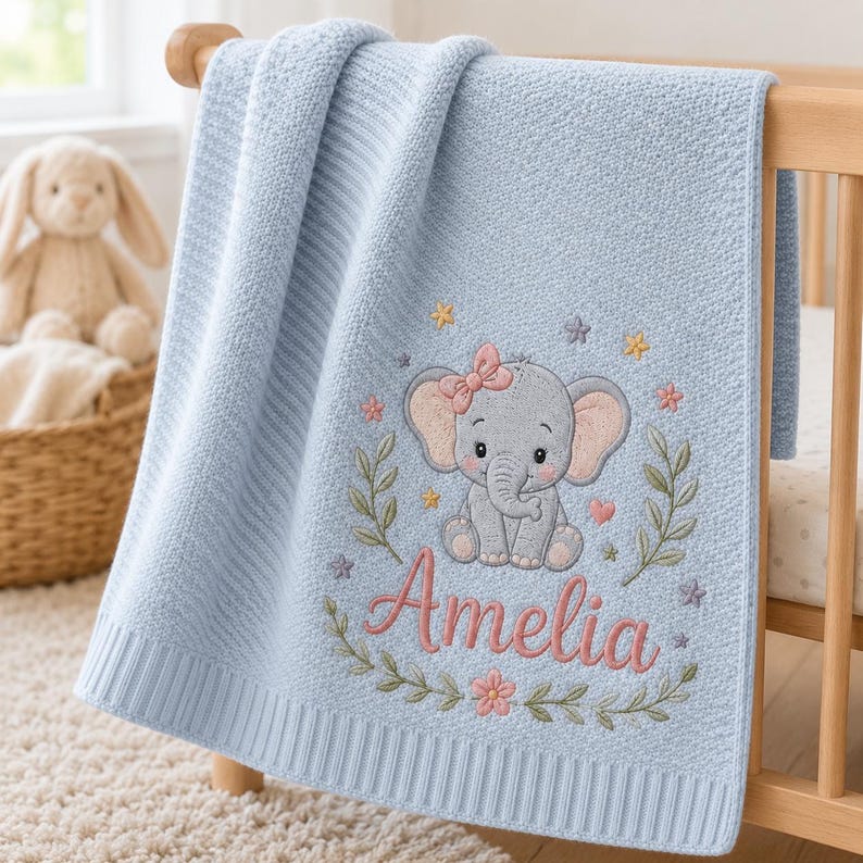 May include: A light blue knitted baby blanket featuring an embroidered gray elephant with a pink bow, surrounded by floral vines, small stars, and hearts. The name Amelia is embroidered in pink script lettering below the elephant design.