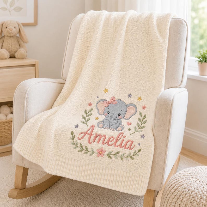 May include: A cream-colored knit blanket featuring an embroidered design of a gray elephant with a pink bow, surrounded by green leafy branches, small colorful flowers, and stars. The name Amelia is embroidered in pink script below the ele