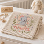 May include: A beige knitted baby blanket featuring an embroidered gray elephant with a pink bow, surrounded by small colorful flowers and green leaves. The name Amelia is embroidered in pink script below the elephant. The blanket has a rib