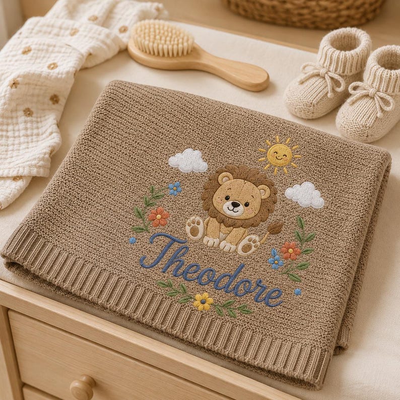 May include: A tan knitted baby blanket featuring an embroidered lion, a yellow sun, white clouds, and colorful flowers. The name Theodore is embroidered in blue script below the lion. Nearby are a wooden hairbrush and cream-colored booties