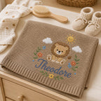 May include: A tan knitted baby blanket featuring an embroidered lion, a yellow sun, white clouds, and colorful flowers. The name Theodore is embroidered in blue script below the lion. Nearby are a wooden hairbrush and cream-colored booties