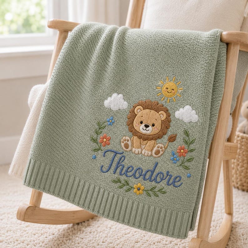 May include: A sage green knitted baby blanket featuring an embroidered design of a brown lion, a yellow sun, white clouds, and colorful flowers. The name Theodore is embroidered in blue cursive script below the lion, with a ribbed border.
