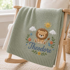 May include: A sage green knitted baby blanket featuring an embroidered design of a brown lion, a yellow sun, white clouds, and colorful flowers. The name Theodore is embroidered in blue cursive script below the lion, with a ribbed border.