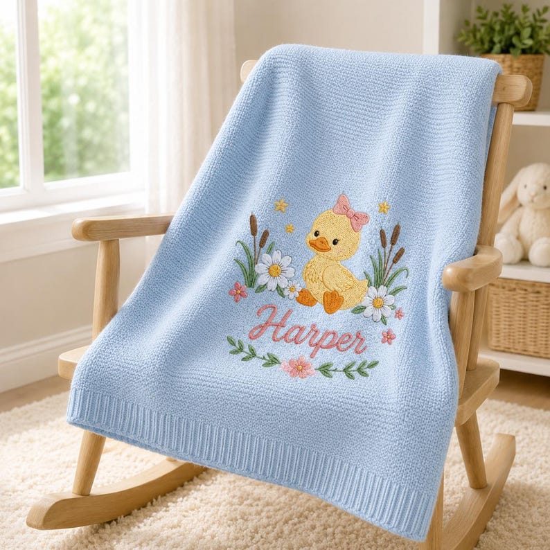 May include: A light blue knitted baby blanket featuring an embroidered design of a yellow duckling with a pink bow, surrounded by white daisies, green reeds, and small yellow stars. The name Harper is embroidered in pink cursive script bel