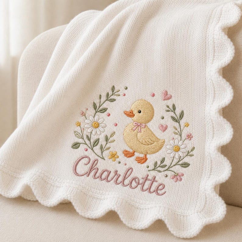 May include: A cream-colored knit blanket with a scalloped edge featuring an embroidered design of a yellow duckling surrounded by green leaves, white daisies, and small pink hearts, with the name Charlotte stitched in pink cursive script.