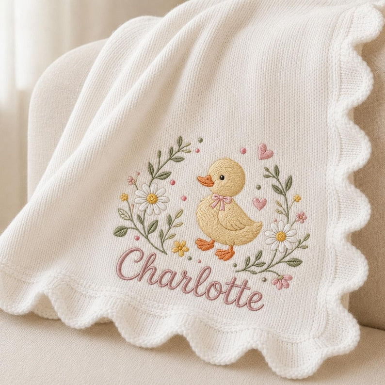 May include: A cream-colored knit blanket with a scalloped edge featuring an embroidered design of a yellow duckling surrounded by green leaves, white daisies, and small pink hearts, with the name Charlotte stitched in pink cursive script.
