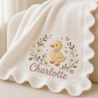 May include: A cream-colored knit blanket with a scalloped edge featuring an embroidered design of a yellow duckling surrounded by green leaves, white daisies, and small pink hearts, with the name Charlotte stitched in pink cursive script.