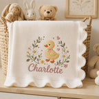 May include: A cream-colored knit baby blanket with a scalloped edge, featuring an embroidered design of a yellow duckling surrounded by daisies, green leaves, and small pink hearts, with the name Charlotte stitched in pink cursive script below.