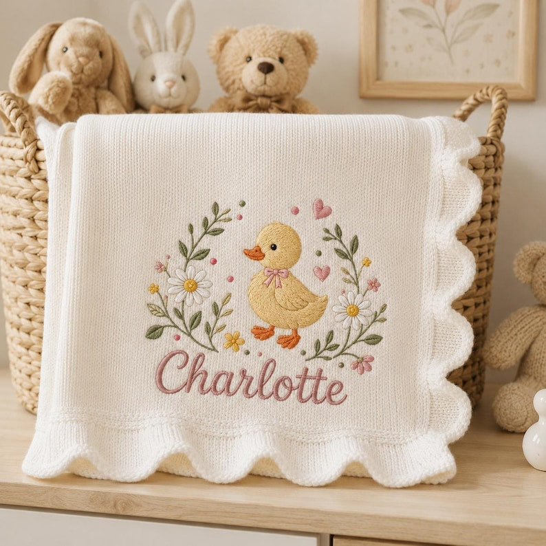 May include: A cream-colored knit baby blanket with a scalloped edge, featuring an embroidered design of a yellow duckling surrounded by daisies, green leaves, and small pink hearts, with the name Charlotte stitched in pink cursive script b