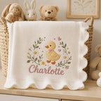 May include: A cream-colored knit baby blanket with a scalloped edge, featuring an embroidered design of a yellow duckling surrounded by daisies, green leaves, and small pink hearts, with the name Charlotte stitched in pink cursive script b