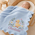 May include: A light blue knitted baby blanket with a scalloped edge, featuring an embroidered design of a yellow duckling, white daisies, green leaves, and small pink hearts, with the name Charlotte stitched in pink cursive script lettering.