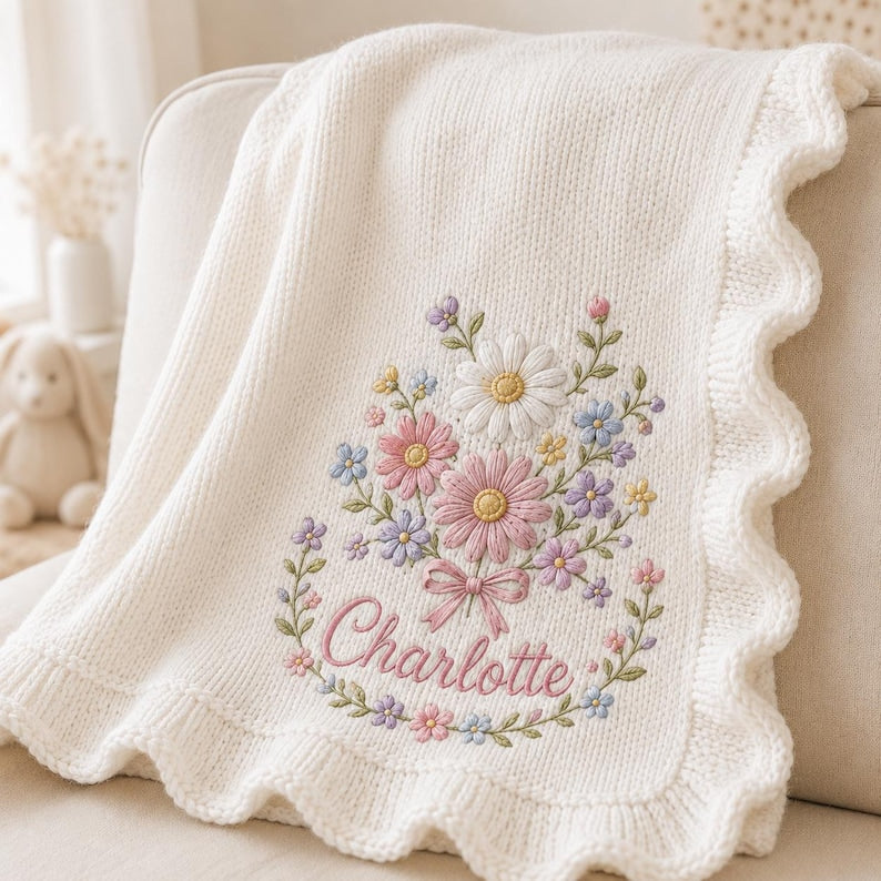 May include: A cream-colored knitted blanket featuring a scalloped edge and an embroidered floral bouquet design. The center of the embroidery includes the name Charlotte in pink script, surrounded by daisies and small flowers in pastel col