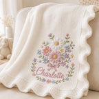 May include: A cream-colored knitted blanket featuring a scalloped edge and an embroidered floral bouquet design. The center of the embroidery includes the name Charlotte in pink script, surrounded by daisies and small flowers in pastel col