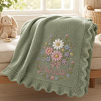 May include: A sage green knitted baby blanket featuring a scalloped edge and an embroidered floral bouquet design. The center of the blanket displays a cluster of daisies and small flowers in pink, white, and blue, with the name Charlotte 