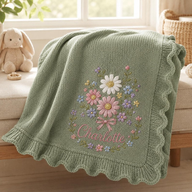 May include: A sage green knitted baby blanket featuring a scalloped edge and an embroidered floral bouquet design. The center of the blanket displays a cluster of daisies and small flowers in pink, white, and blue, with the name Charlotte 