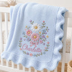 May include: A light blue knitted baby blanket with a ruffled edge, featuring an embroidered floral bouquet in shades of pink, white, yellow, and purple, with the name Charlotte stitched in pink cursive script below the flowers.