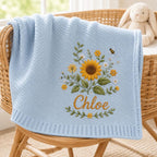 May include: A light blue knit baby blanket featuring an embroidered bouquet of yellow sunflowers, white daisies, and green leaves. A small bee is embroidered above the flowers. The name Chloe is embroidered in gold script below the floral 
