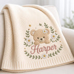 May include: A cream-colored knit blanket featuring an embroidered design of a light brown teddy bear surrounded by a wreath of green leaves, small yellow stars, and white daisies. The name Harper is embroidered in pink cursive script below.