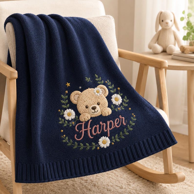 May include: A navy blue knitted blanket featuring an embroidered design of a light brown teddy bear surrounded by white daisies, green leaves, and small yellow stars. The name Harper is embroidered in pink cursive script below the bear.