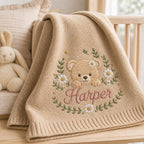 May include: A beige knitted baby blanket featuring an embroidered design of a teddy bear surrounded by a wreath of green leaves and white daisy flowers. The name Harper is embroidered in pink cursive script below the bear's paws.