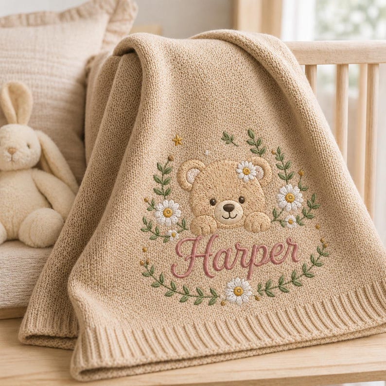 May include: A beige knitted baby blanket featuring an embroidered design of a teddy bear surrounded by a wreath of green leaves and white daisy flowers. The name Harper is embroidered in pink cursive script below the bear's paws.