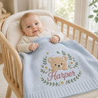 May include: A light blue knitted baby blanket featuring an embroidered tan teddy bear surrounded by a wreath of green leaves and white daisies. The name Harper is stitched in pink cursive script below the bear. The blanket has a ribbed border.