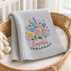 May include: A light gray knit blanket featuring an embroidered bouquet of colorful flowers including pink, blue, yellow, and purple blossoms with green stems and leaves. The name Sophie is embroidered in pink script below the floral arrang