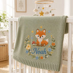 May include: A sage green knitted baby blanket featuring an embroidered orange and white fox sitting next to a red mushroom. The design includes a floral wreath with blue, yellow, and orange flowers, and the name Noah in blue script letteri