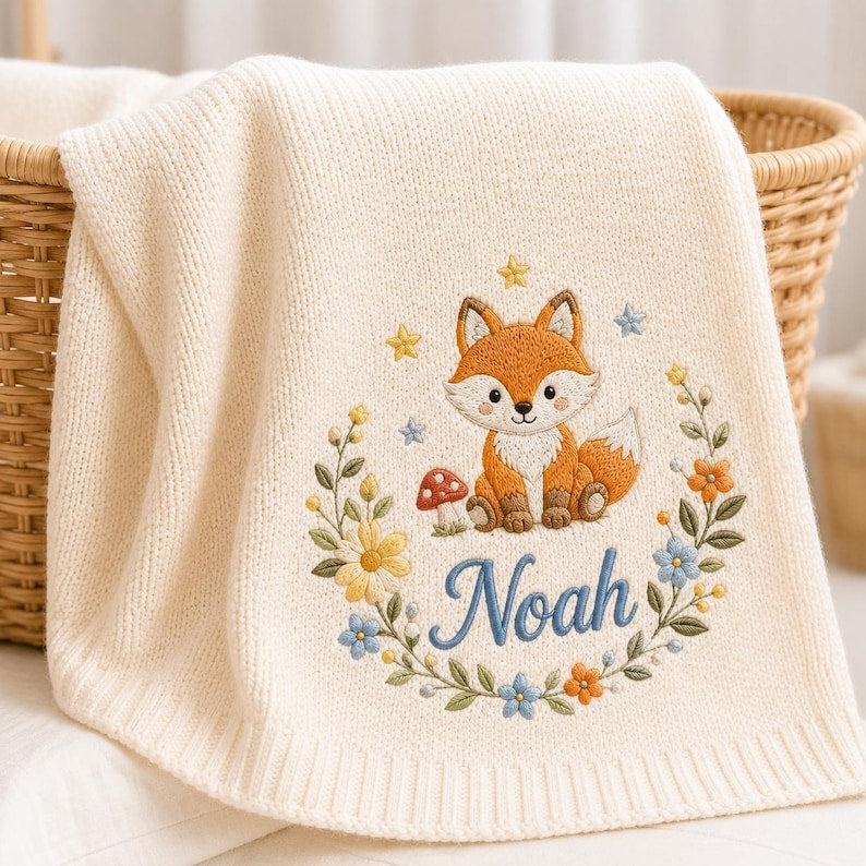 May include: A cream-colored knit blanket featuring an embroidered design of a fox sitting next to a red mushroom. The fox is surrounded by a floral wreath with yellow, orange, and blue flowers, small stars, and the name Noah stitched in bl