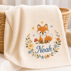 May include: A cream-colored knit blanket featuring an embroidered design of a fox sitting next to a red mushroom. The fox is surrounded by a floral wreath with yellow, orange, and blue flowers, small stars, and the name Noah stitched in bl