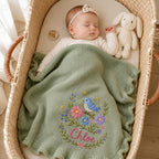 May include: A sage green knitted baby blanket with a ruffled edge, featuring an embroidered design of a blue bird perched on a branch surrounded by pink and purple flowers, with the name Chloe stitched in pink cursive script at the bottom.