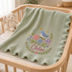 May include: A sage green knitted baby blanket with a scalloped edge, featuring an embroidered design of a blue bird perched on a branch surrounded by pink, blue, and purple flowers, with the name Chloe stitched in pink cursive lettering be