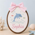 May include: An oval embroidery hoop featuring a blue dolphin, small fish, and bubbles stitched on white fabric. The name Sophia is embroidered in pink script below the dolphin. A pink and white gingham bow is attached to the top of the woo