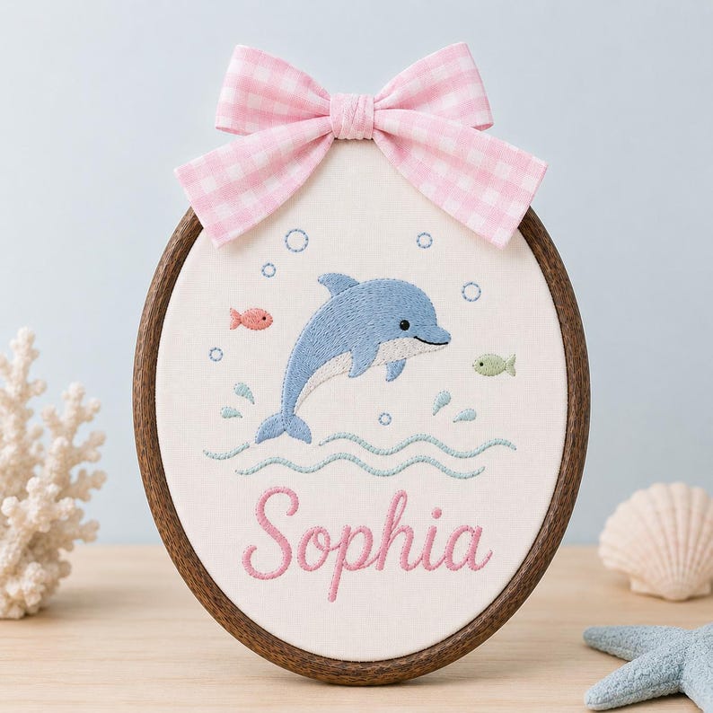 May include: An oval embroidery hoop featuring a blue dolphin, small fish, and bubbles stitched on white fabric. The name Sophia is embroidered in pink script below the dolphin. A pink and white gingham bow is attached to the top of the woo