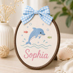 May include: An oval embroidery hoop frame containing a cream-colored fabric panel with a blue dolphin, small fish, and bubbles stitched in the center. The name Sophia is embroidered in pink script below, topped with a blue and white gingha