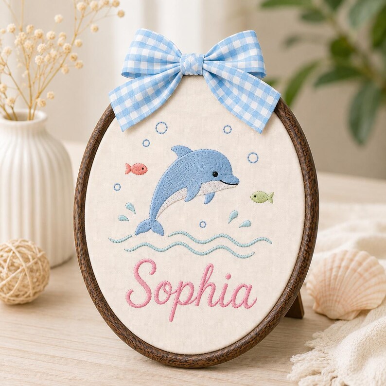 May include: An oval embroidery hoop frame containing a cream-colored fabric panel with a blue dolphin, small fish, and bubbles stitched in the center. The name Sophia is embroidered in pink script below, topped with a blue and white gingha