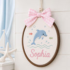 May include: An oval embroidery hoop wall hanging featuring a blue dolphin, small fish, and bubbles stitched on white fabric. The name Sophia is embroidered in pink script below the dolphin. A pink and white gingham bow is attached to the t
