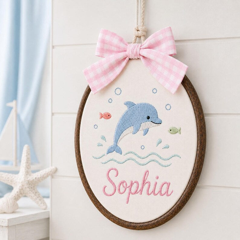 May include: An oval embroidery hoop wall hanging featuring a blue dolphin, small fish, and bubbles stitched on white fabric. The name Sophia is embroidered in pink script below the dolphin. A pink and white gingham bow is attached to the t