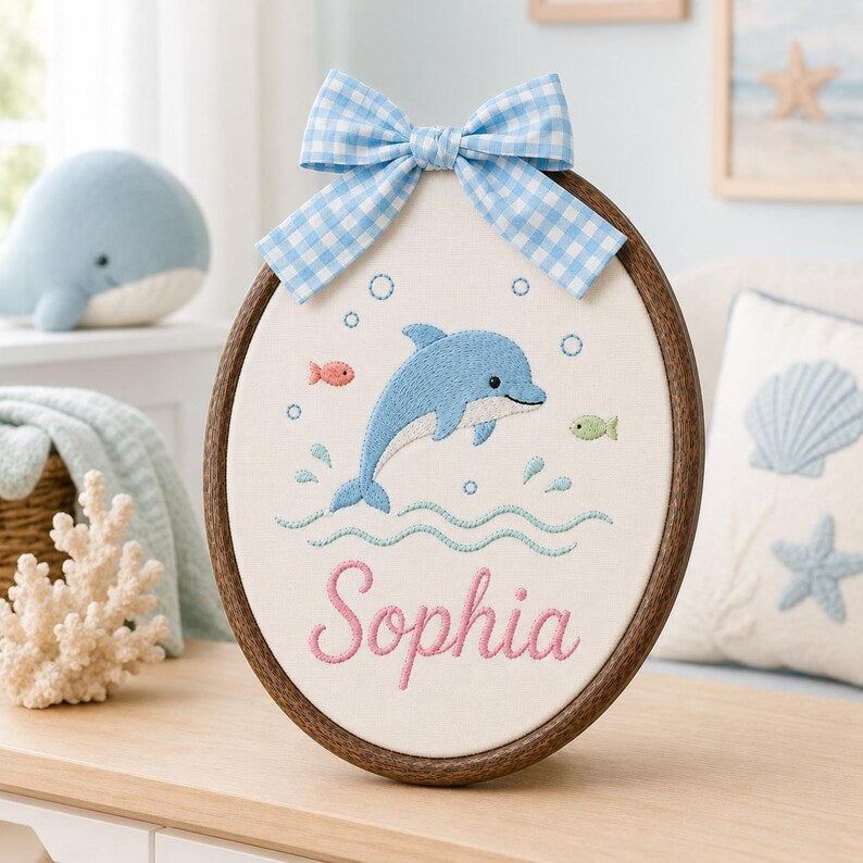 May include: An oval embroidery hoop wall hanging featuring a blue dolphin, small fish, and bubbles stitched on white fabric. The name Sophia is embroidered in pink script below the dolphin. A blue and white gingham ribbon bow is attached a