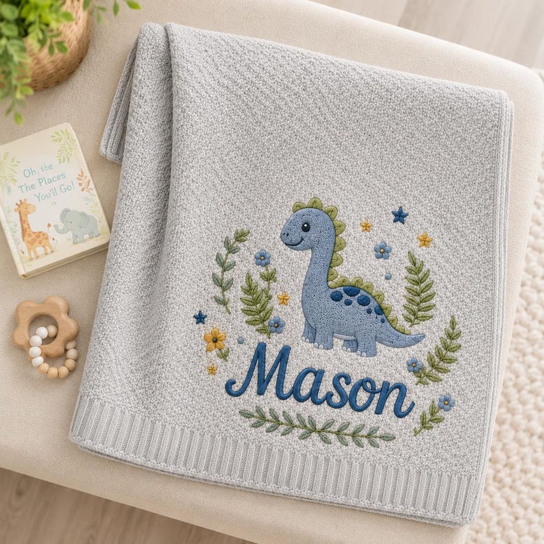 May include: A light gray knit blanket featuring an embroidered blue dinosaur surrounded by green foliage and small flowers. The name Mason is embroidered in dark blue script below the dinosaur. The blanket has a ribbed border along the edge.