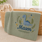 May include: A sage green knitted baby blanket featuring an embroidered blue dinosaur surrounded by small flowers, leaves, and stars. The name Mason is embroidered in dark blue script below the dinosaur. The blanket has a ribbed border edge.