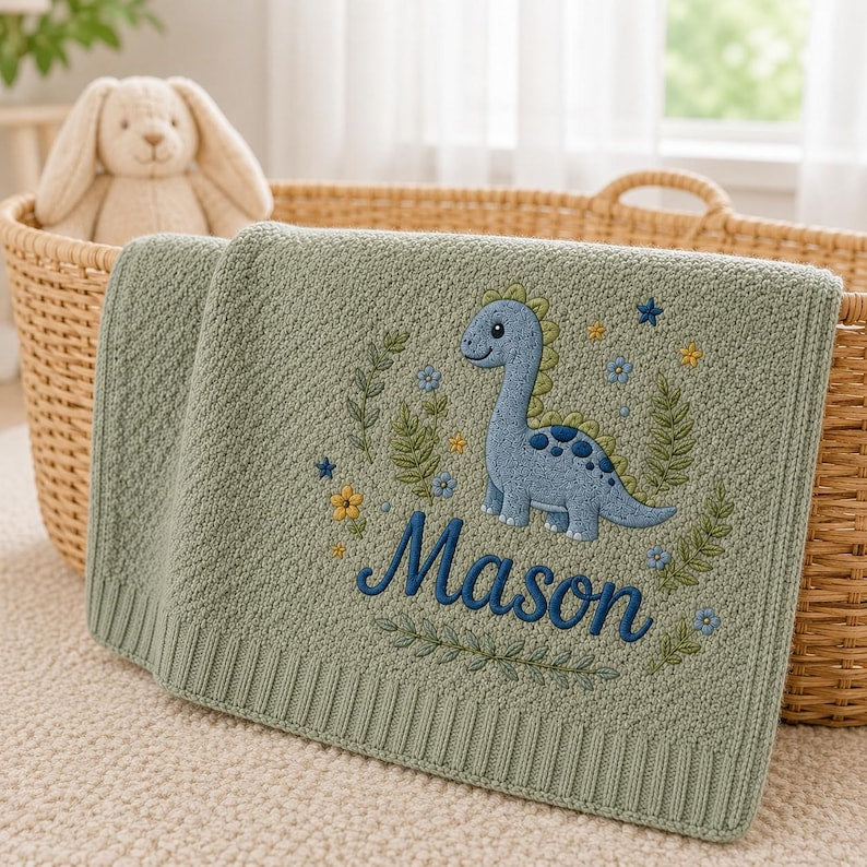 May include: A sage green knitted baby blanket featuring an embroidered blue dinosaur surrounded by small flowers, leaves, and stars. The name Mason is embroidered in dark blue script below the dinosaur. The blanket has a ribbed border edge