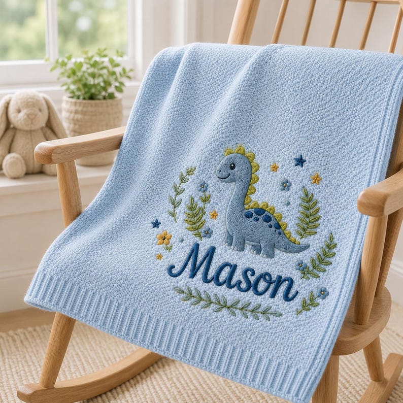 May include: A light blue knit baby blanket featuring an embroidered design of a blue dinosaur surrounded by green leaves, small yellow and blue flowers, and stars. The name Mason is embroidered in dark blue script lettering below the dinosaur.