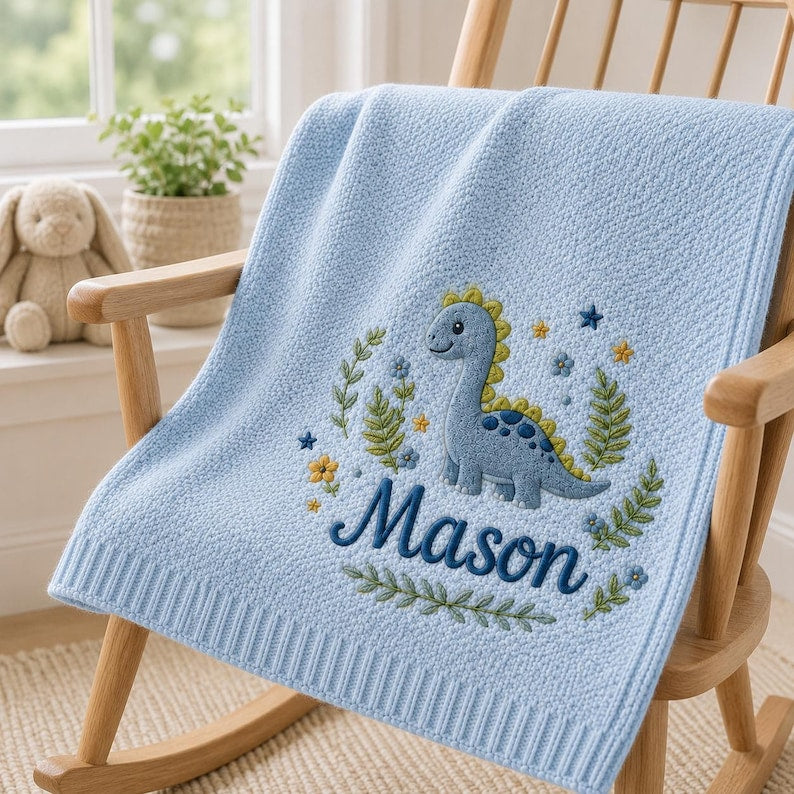 May include: A light blue knit baby blanket featuring an embroidered design of a blue dinosaur surrounded by green leaves, small yellow and blue flowers, and stars. The name Mason is embroidered in dark blue script lettering below the dinos